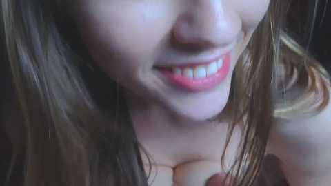 Media: A close-up video of a young woman with fair skin, long brown hair, and red lipstick, smiling broadly, revealing white teeth. The image is out of focus, emphasizing her face.