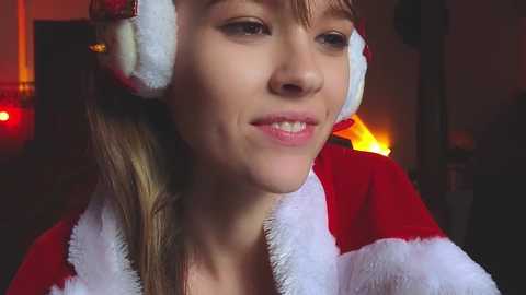 Media: Video of a young Caucasian woman with light brown hair, wearing a Santa hat and a red fuzzy robe, smiling softly, in a warmly lit, softly blurred background.