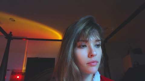 Media: Video of a young woman with shoulder-length blonde hair, wearing a red top, in a dimly lit bedroom with warm lighting, slightly out of focus.