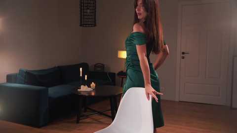 Media: Video of a curvy woman in a green off-shoulder dress, standing in a dimly lit living room with wooden floor, blue couch, and modern decor.