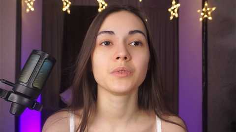 Media: Video of a young woman with long, straight brown hair, wearing a white tank top, making a pouty face into a microphone. Background features fairy lights and a dark wooden door.