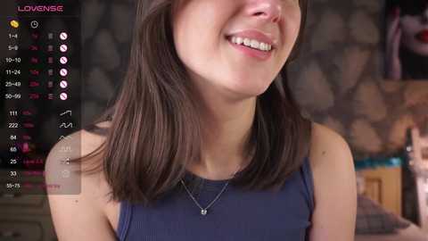 Video of a smiling woman with straight, shoulder-length brown hair, wearing a sleeveless dark blue top, standing in a dimly lit room with a stone wall background.