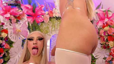 Media: Video of a blonde drag queen with long hair and heavy makeup, tongue out, in a pink, floral background.