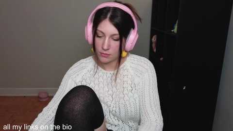 Media: Video of a young woman with fair skin, dark hair, and a white knitted sweater, wearing pink headphones and black tights, sitting on a wooden floor, looking down, in a dimly lit room with a bookshelf in the background.