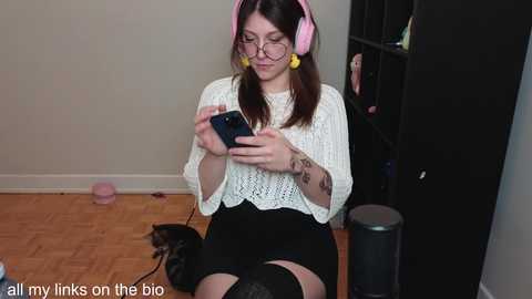 Media: Video of a young woman with glasses, pink headphones, and a white knit top, sitting on a wooden floor, focused on her smartphone, surrounded by a cluttered room with a cat and a black shelf.