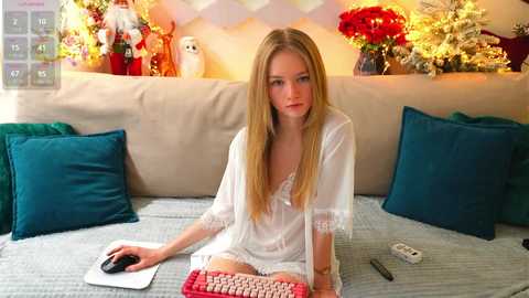 Media: Video of a blonde, fair-skinned young woman in a sheer white nightgown, sitting on a beige couch with teal pillows. Background features Christmas decorations and a mousepad.