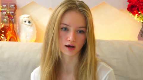 Media: Video of a young, fair-skinned blonde woman with blue eyes, wearing a white top, sitting on a beige couch. Background features a white owl statue and a vase of red roses.