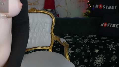 Media: Video of a person with a large breast, dressed in a black jacket, sitting on a gold-framed white chair in a room with a black and white snowflake-patterned blanket, a Christmas tree, and a red and gold wall.