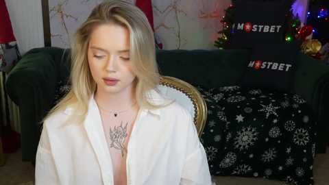 Media: Video of a blonde young woman with fair skin, wearing a white shirt and black necklace, sitting on a sofa with holiday decorations in the background.