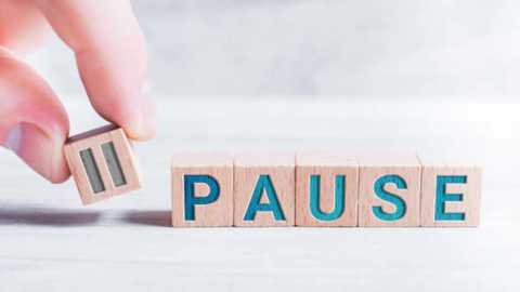 Media: Video of a light-skinned hand holding a wooden block with the letter \"A\" in blue, placing it on a stack of wooden blocks spelling \"PAUSE\". The background is a blurred, light gray wall.