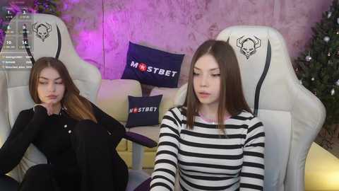 Video of two young women in gaming chairs, one with straight brown hair, one with long brown hair, both wearing black and white striped shirts, in a cozy, dimly-lit room with purple lighting and a decorated Christmas tree.