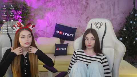 Video of two young women in a cozy living room. One wears a red Santa hat, the other a striped shirt. Both sit on a white gaming chair, surrounded by Christmas decor and purple lighting.