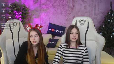 Media: Video of two young women with fair skin, long hair, and festive red headbands, seated in white gaming chairs, wearing casual clothing. Background includes a purple wall, a Christmas tree, and \"MISTREET\" pillow.