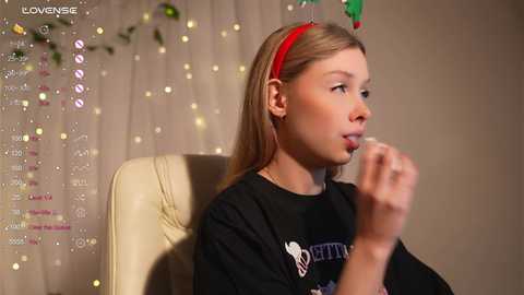 Video of a young girl with light skin and straight blonde hair wearing a red headband, sitting in a white chair. She's eating a cookie, with a festive background of string lights and greenery.