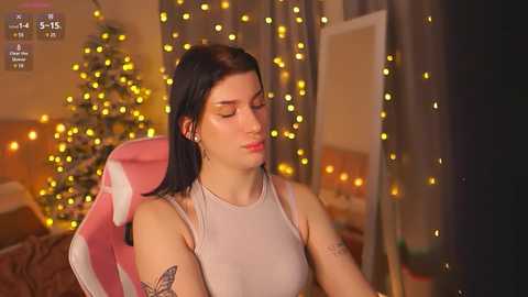 Media: Video of a fair-skinned woman with straight black hair, wearing a light gray tank top, seated in a pink gaming chair, surrounded by warm yellow fairy lights and a decorated Christmas tree in a cozy indoor setting.