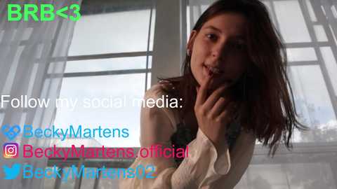 Media: Video of a young woman with shoulder-length brown hair, wearing a white blouse, posing by a window. Text overlays with social media handles and a watermark.