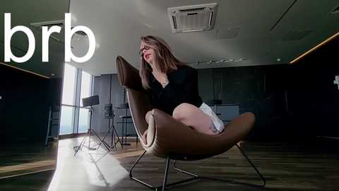 Media: Video of a young woman with long brown hair, wearing a black top and white shorts, sitting on a modern, tan leather chair in a dimly lit room with dark walls and wooden floor.