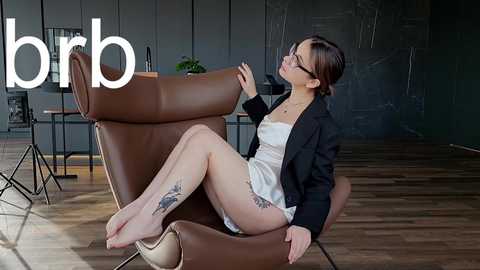 Media: Video of a young Asian woman in a white dress, black blazer, and glasses, reclining in a brown leather chair, with tattoos on her legs, in a modern, minimalist room with wooden floors and dark walls.