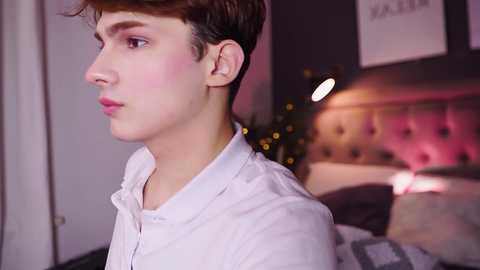 Media: Video of a young, fair-skinned, short-haired man with rosy cheeks, wearing a white shirt, looking contemplative, in a softly lit, modern bedroom with a bed and dimly lit Christmas tree.