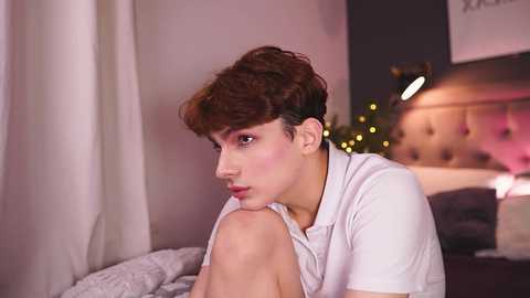 Media: Video of a pale-skinned, androgynous person with short, curly brown hair, wearing a black headband, white shirt, and light makeup, sitting on a bed with a grey blanket, in a softly lit bedroom with a tufted headboard and string lights.