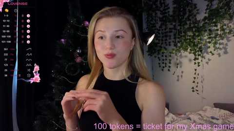 Media: A video of a young woman with long blonde hair, fair skin, and a slender build, wearing a black sleeveless top, smiling. Background includes a dark room with greenery and a bed with a white blanket. Text reads \"1000 tokens = tickled for my Xmas gift\" in pink.