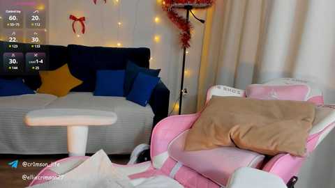 Media: Video of a cozy living room with a dark blue couch, yellow and blue pillows, a white chair, and a pink gaming chair, adorned with festive string lights and a small Christmas tree.