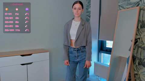 Media: Video of a young Caucasian woman with light skin, brown hair, wearing a grey blazer, white crop top, and high-waisted jeans, standing in a modern, minimalist room with a large mirror and digital clock on the wall.