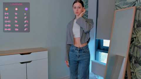 Media: A video of a young woman with fair skin and straight brown hair, wearing a gray cropped jacket over a white crop top and high-waisted blue jeans, posing in a modern, minimalist room with a light green wall and a digital clock displaying \"1:12\".