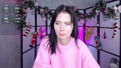 Media: Video of a fair-skinned, black-haired woman with a neutral expression wearing a pink sweater, surrounded by festive holiday decorations, including garlands and Christmas stockings, in a dimly lit room.