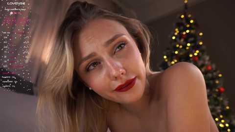 Media: Video of a blonde woman with fair skin, red lipstick, and bare shoulders, looking contemplative. Background includes a festive Christmas tree with golden lights.