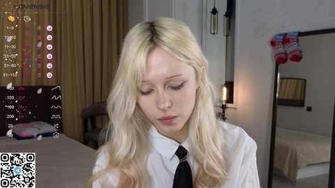 Video of a young, fair-skinned, blonde woman in a white shirt and black tie, with a neutral expression, in a modern bedroom with beige curtains, a bed, and a lamp.