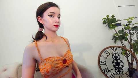 Media: A video of a young Caucasian woman with fair skin and a slender build, wearing a floral orange crop top, sitting indoors. She has a pouty red lipstick and a single braid. Background includes a green plant and a vintage-style clock.