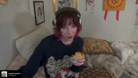 Media: A video of a fair-skinned woman with shoulder-length brown hair, wearing a black hoodie and headphones, sitting on a bed with a leopard-print blanket. She holds a sandwich. The room has a cozy, cluttered feel with various knick-knacks and decorations on the walls.