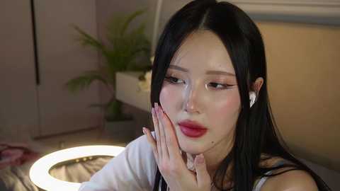 Media: Video of an East Asian woman with long black hair, wearing red lipstick, and touching her face. She's in a modern, softly lit room with a potted plant and a round mirror in the background.