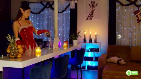 Media: Video of a woman in a red Santa outfit, elf ears, and red gloves, mixing drinks at a bar counter with a pineapple and candles. Background: modern living room with cozy ambiance, dim lighting, and Christmas decor.