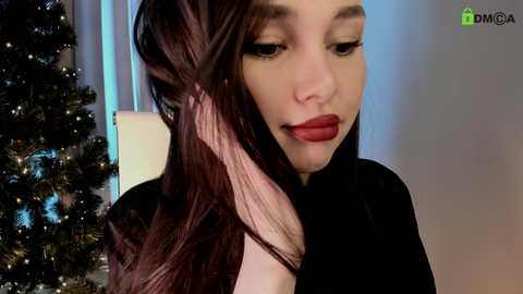 Video of a woman with long, wavy, dark brown hair, wearing black clothing and bold red lipstick. She is brushing her hair and looking down. Background includes a lit Christmas tree and soft, blue lighting.