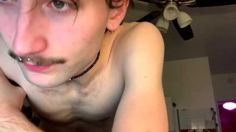 Media: Video of a young, shirtless man with fair skin, blue eyes, and a trimmed mustache, wearing a black choker. He is in a dimly lit room with a ceiling fan and a blurred background.