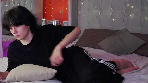 Media: A video of a young man with messy black hair and a black t-shirt lying on a bed with beige pillows and a tufted headboard. He's wearing black Adidas shorts.