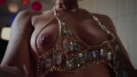 Media: A close-up video of a topless woman with fair skin, wearing a gold chain necklace with large, faceted gemstones. Her breasts are partially covered by the necklace, revealing her nipples. The background is blurred with bokeh lights.