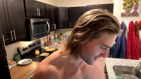 Media: Video of a shirtless man with long brown hair, cooking in a modern kitchen with dark wood cabinets, stainless steel appliances, and red towels hanging.