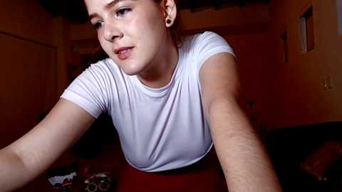 Media: Video of a young, fair-skinned woman with brown hair, wearing a white T-shirt, taking a selfie in a dimly lit room with wooden furniture and muted colors.