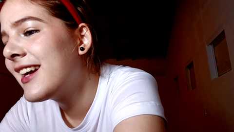 Media: Video of a young woman with light skin, brown hair tied back, wearing a white T-shirt, black ear gauges, and a red headband. She's smiling, looking slightly to the side. Background is dimly lit, with a wall and window visible.