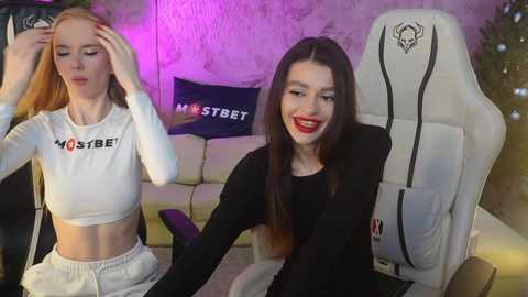 Media: Video of two young women with pale skin, one with long red hair, the other with long dark hair. They are seated in a modern gaming chair, wearing white crop tops, in a cozy living room with purple lights.