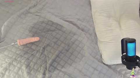 Media: Video of a gray, quilted bedspread with a pink vibrator on top. A black and blue massager is placed on the right side. The image features a minimalist, modern bedroom setting.