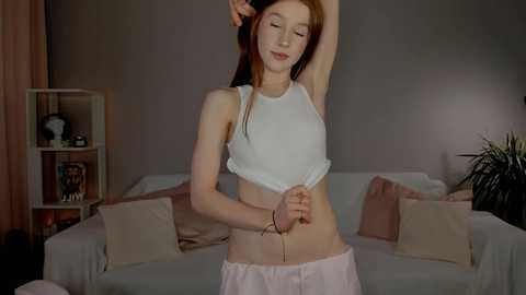 Media: Video of a slender, fair-skinned young woman with red hair, wearing a white crop top and light pink shorts, stretching in a cozy, dimly-lit living room with beige walls, a sofa, and potted plants.