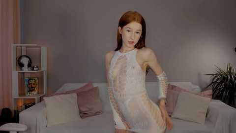 Media: A video of a pale-skinned, red-haired woman in a sheer, floral-patterned dress, standing in a dimly lit living room with a white couch, pink pillows, and a bookshelf.