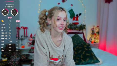 Media: Video of a young woman with curly blonde hair in pigtails, smiling, wearing a beige hoodie, sitting on a bed with Christmas decorations.