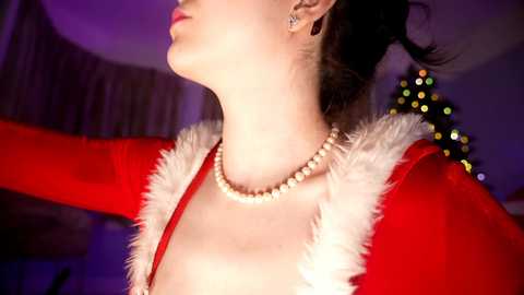 Media: Video of a fair-skinned woman in a red velvet dress with white fur trim, pearl necklace, and earrings, with a Christmas tree and festive lights in the background.