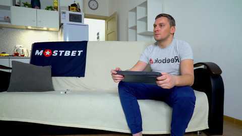 Media: A video of a Caucasian man with short brown hair, wearing a gray t-shirt and blue jeans, playing a video game on a white couch in a modern, minimalist living room with white walls, black shelves, and a blue \"MOSTBET\" pillow.