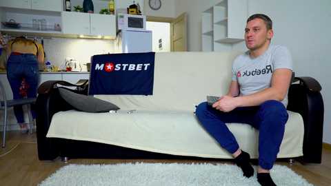 Media: Video of a young man in a gray t-shirt and blue jeans sitting on a white couch in a modern, minimalist living room with a white wall and wooden floor.